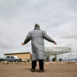 A woman in a gray down winter coat stands with her arms spread in front of a UFO Watchtower tourist attraction.