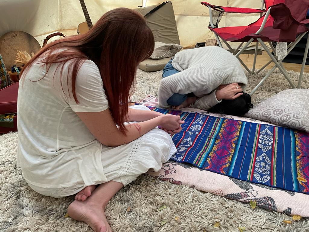A woman sits on the floor with a patient that is holding themselves as they undergo psychedelic-assisted therapy.