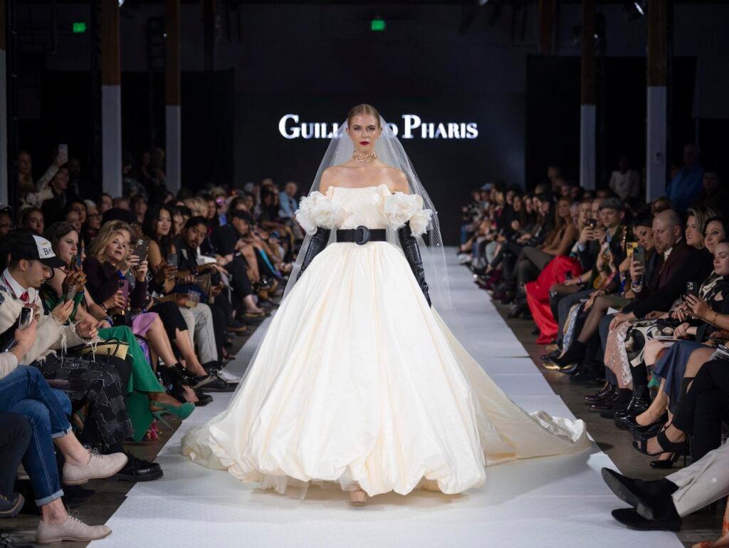 A model in a large, off-white gown with a black belt and tall black gloves walks the runway.