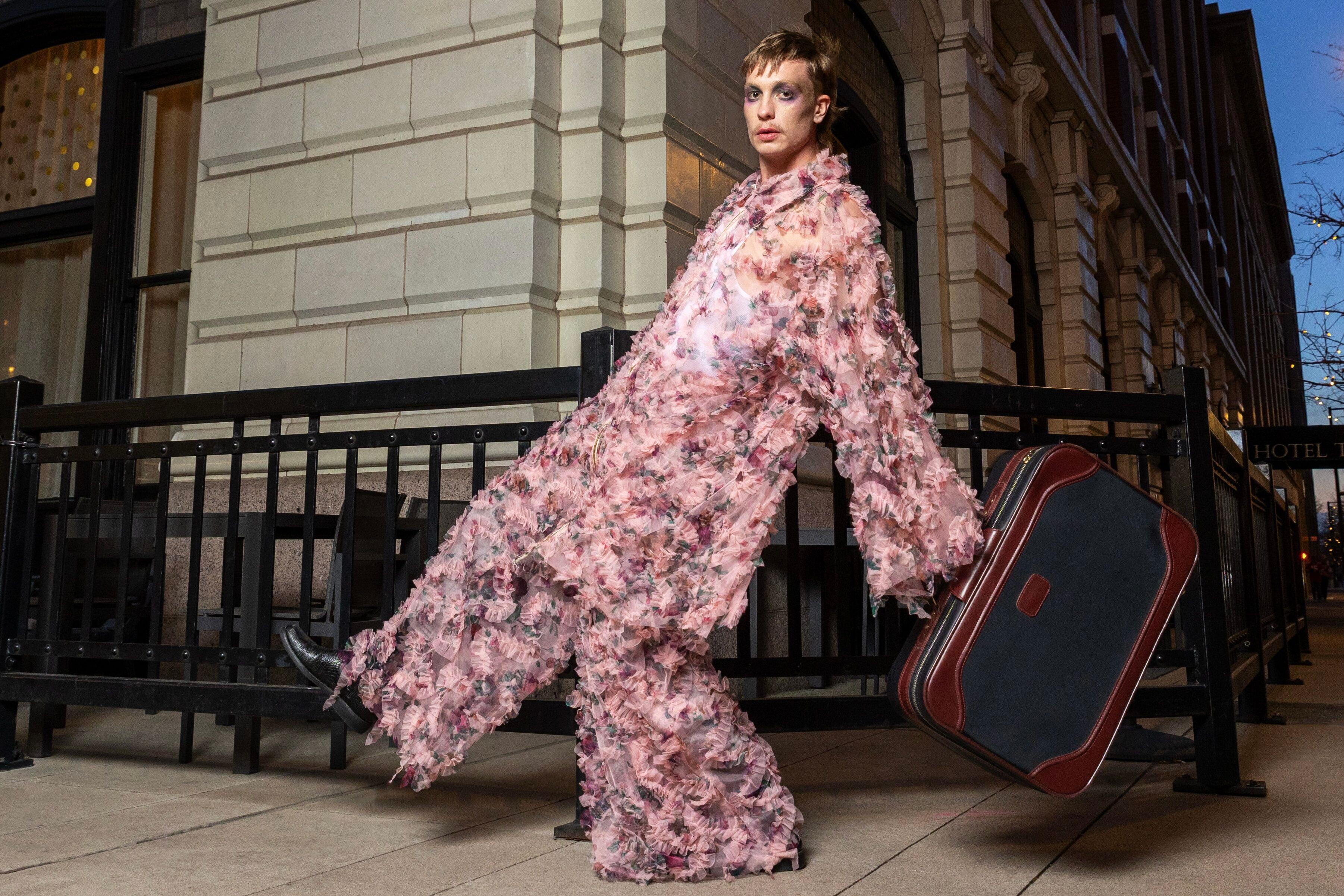 A male model dressed in an oversized pink organza two-piece suit swings a large black and brown suitcase in one arm as he takes an exaggerated step forward.