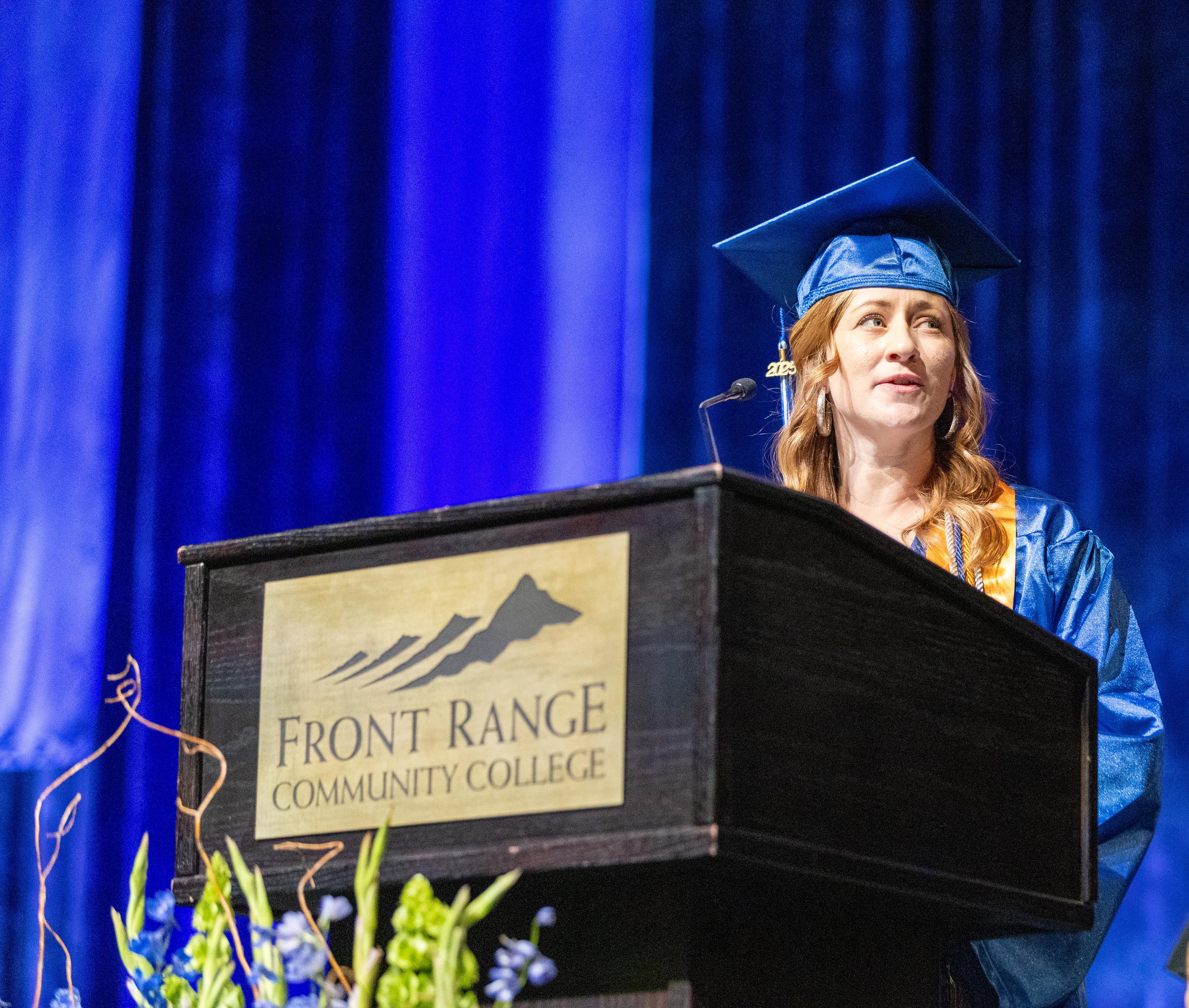 Woman wearing blue graduation cap and gown stands behind a podium in front of a blue backdrop. She is looking to the right.