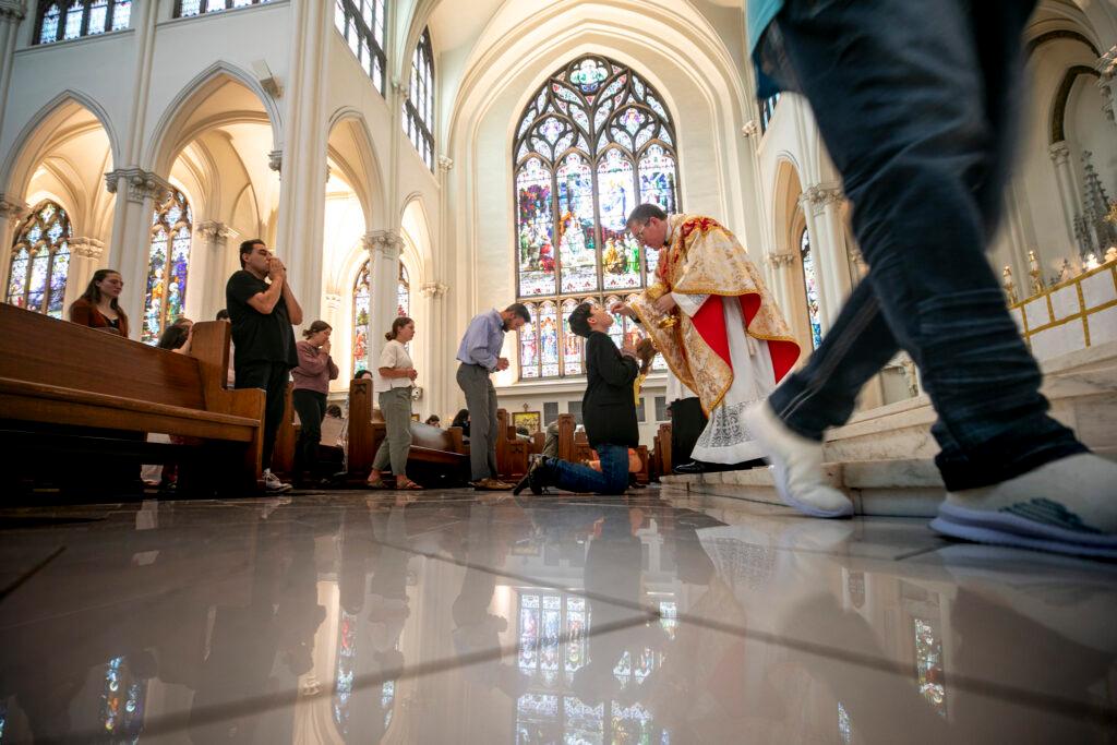 A priest in a robe administers the host to a kneeling person in a large church.
