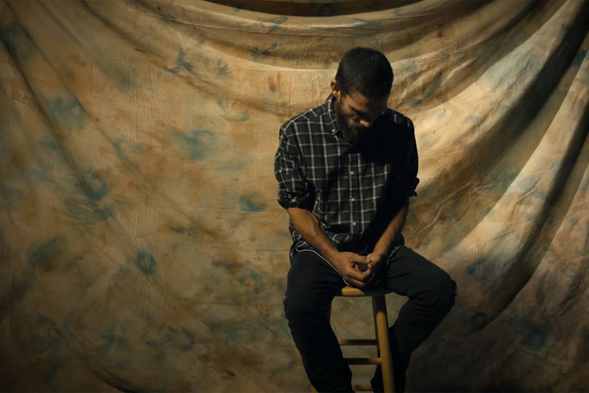 A man sits on a stool looking down in front of a large draped background. He is in shadows.