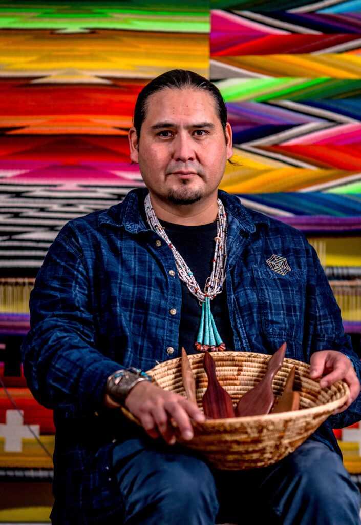 Venacio Aragón sits in front of a rainbow wall of flatweave rugs, holding a basket with wooden tools in it.