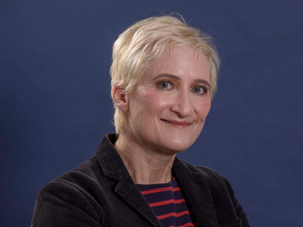 A blonde woman with short hair gazes into the camera in a portrait and a blue background.
