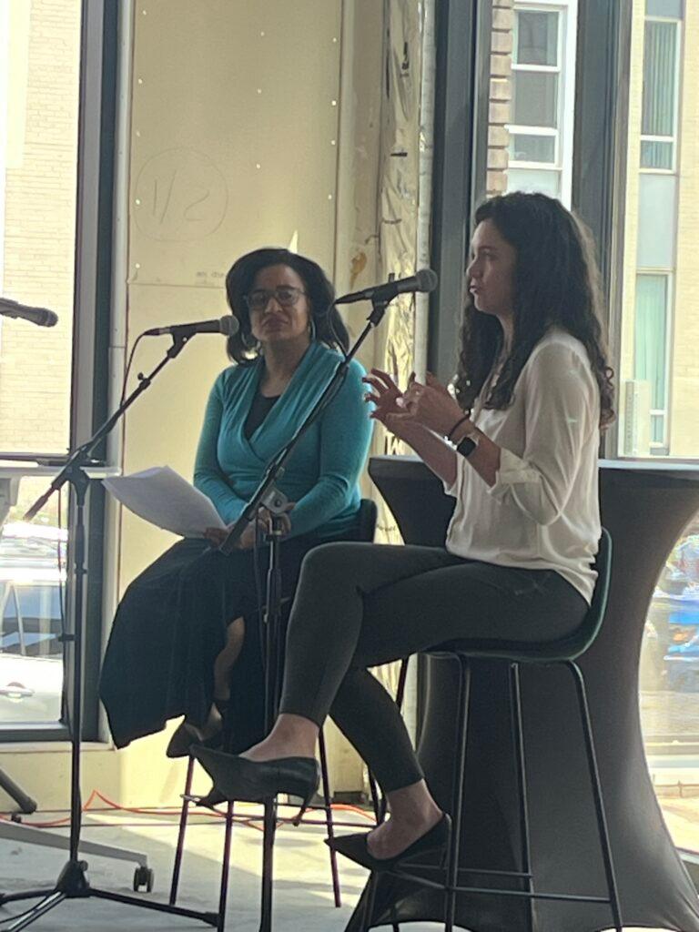Photo shows two women seated on stools with microphones on stands in front of them. The woman on the left, Chandra Thomas Whitfield, is earing a teal top and black skirt and is holding papers. The woman on the right, Kristine Burrows, is wearing a white top with black slacks. They are in front of windows divided by vertical beams.