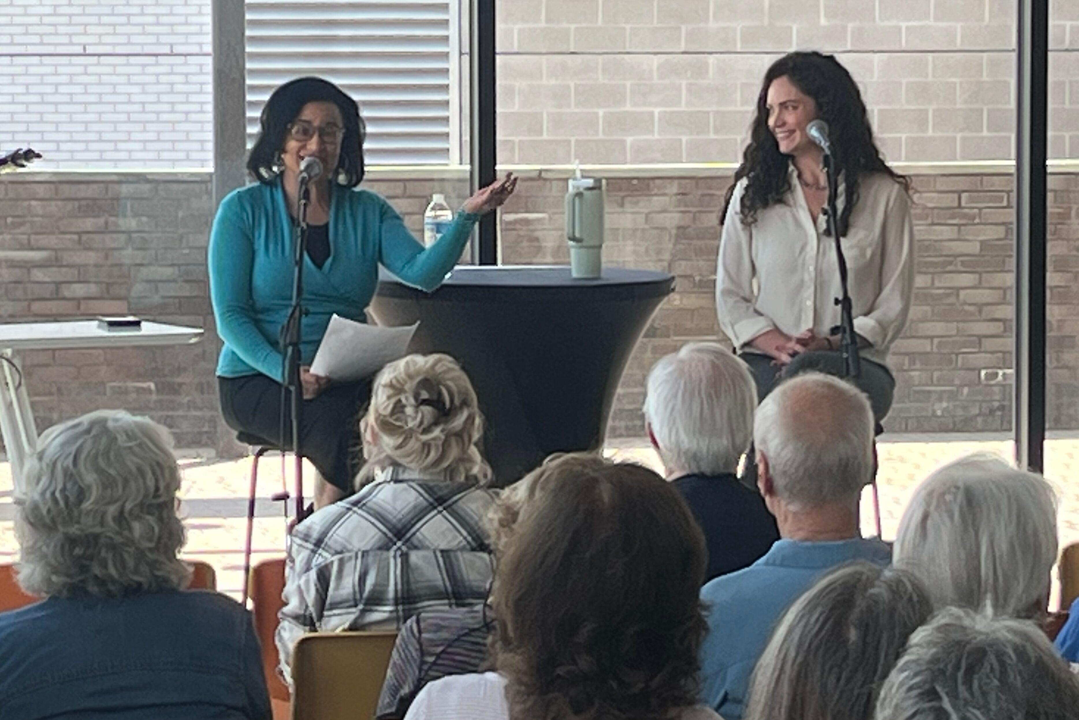 Picture shows two women seated on stools in front of an audience. There is a small black draped table between them. They are seated in front of large floor-to-ceiling windows.
