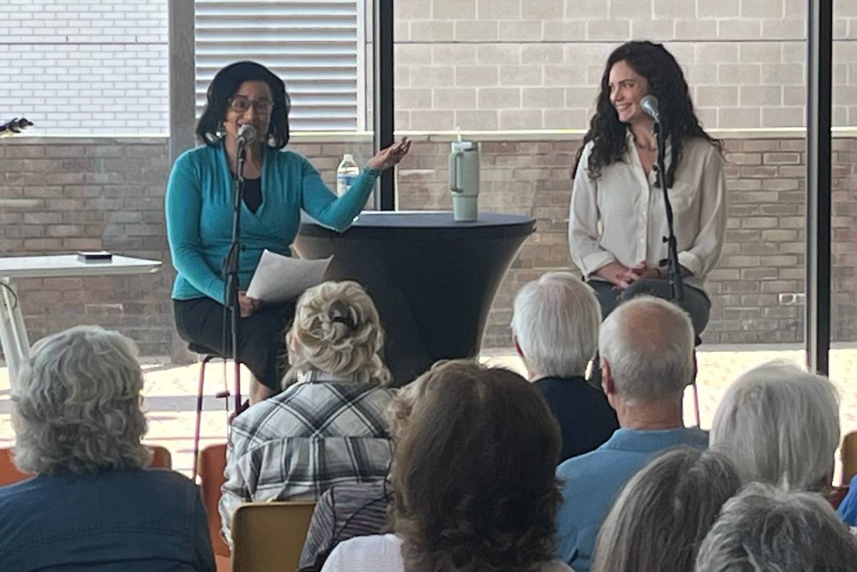 Picture shows two women seated on stools in front of an audience. There is a small black draped table between them. They are seated in front of large floor-to-ceiling windows.