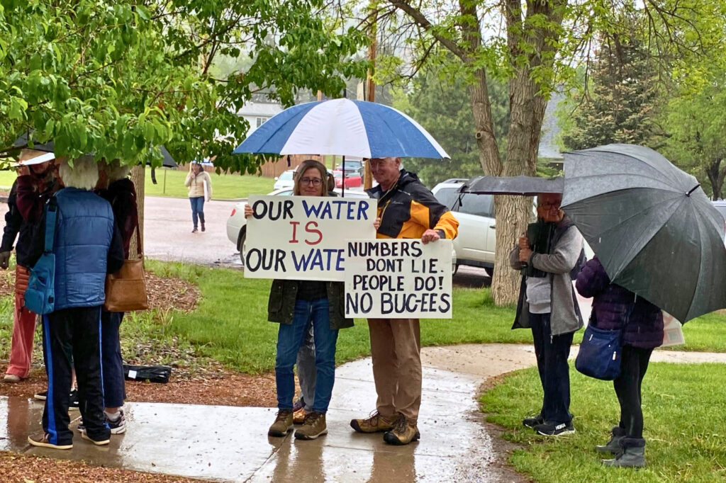 A man and a woman stand under an umbrella with signs that say "Our water is our water," and "Numbers don't lie, people do! No Buc-ees."