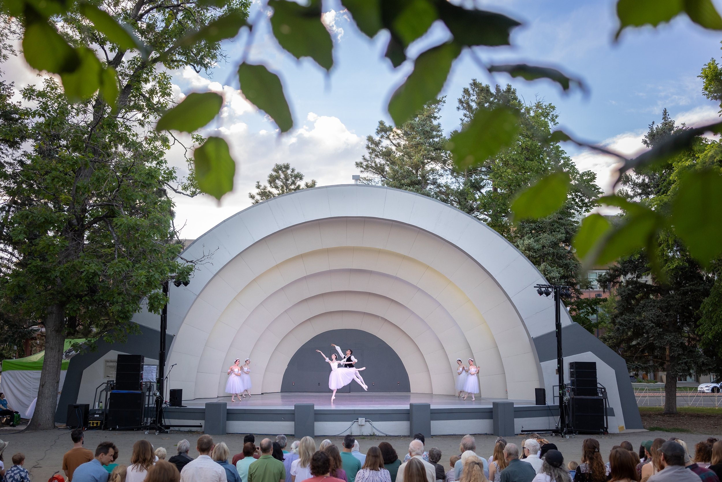 Two ballet dancers leap through the outdoor Bouler Bandshell with a large audeince looking on.
