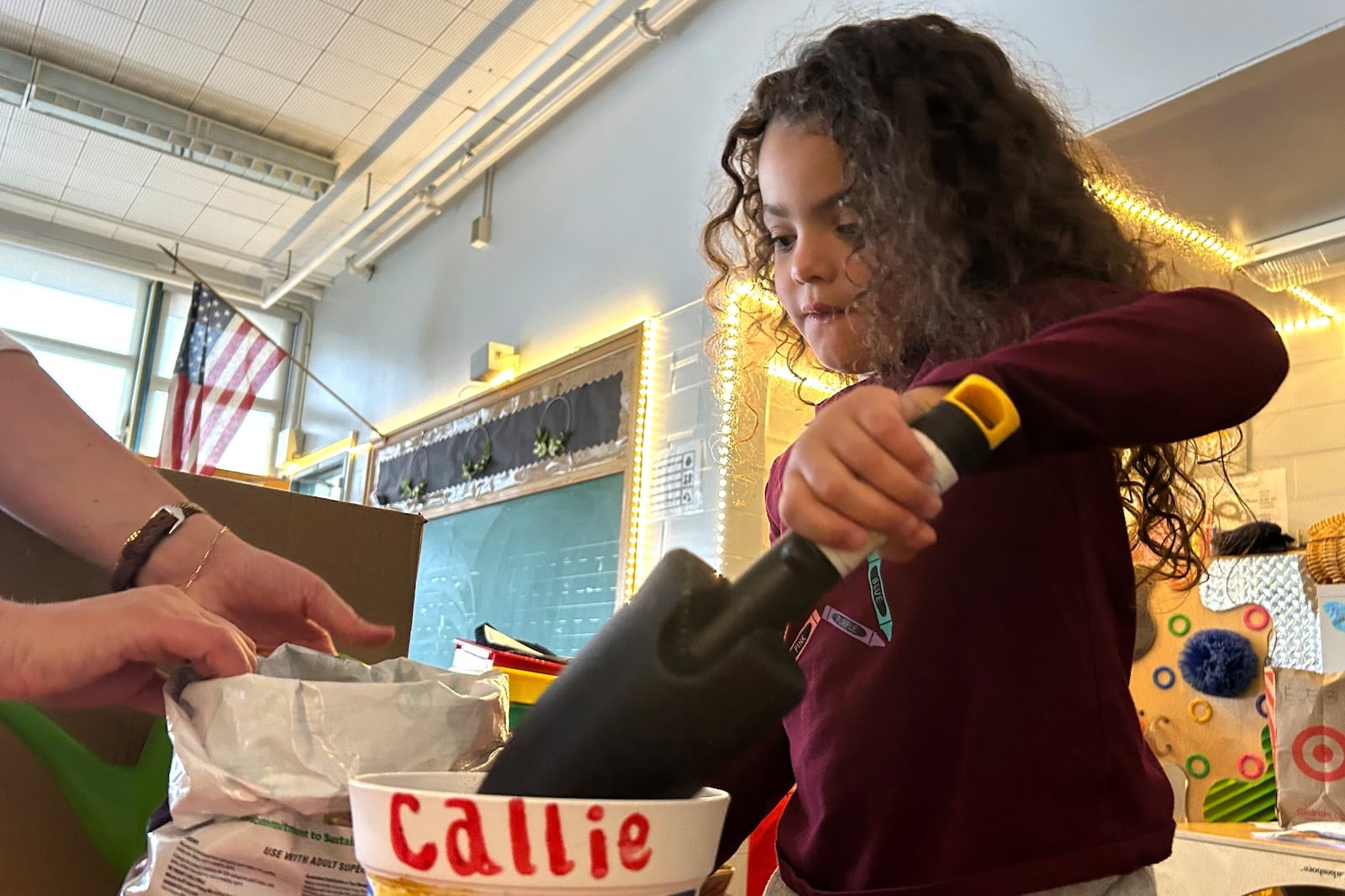 A young girl with curly hair scoops soil into a white pot labeled Callie in red letters. An adult assists nearby. The scene is set in a classroom with an American flag and decorations in the background.
