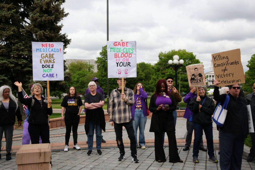 A small crowd of people hold signs on the plaza outside the state capitol