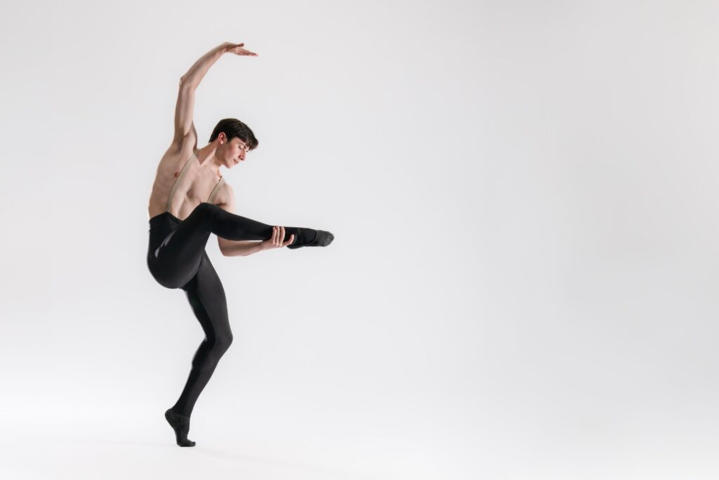 A male dancer poses, right arm arched overhead, right leg grasped, at the ankle, by left hand.
