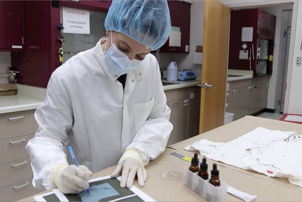 a person in a lab coat, gloves and hair covering writes on a printed sheet