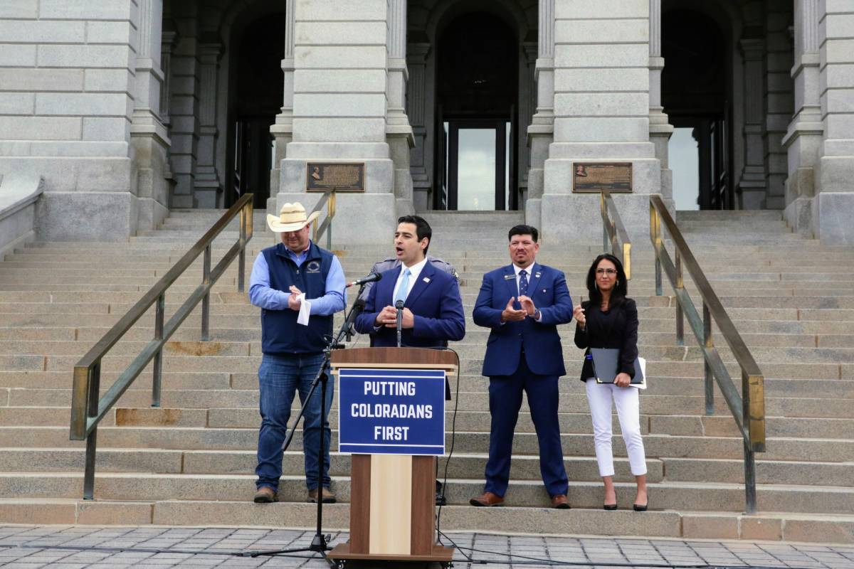 a man in a blue suit stands at a podium with a sign that reads 'putting coloradans first'