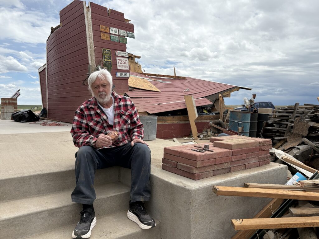 Man with white beard and read plaid shirt sits on porch of destroyed machine shop.