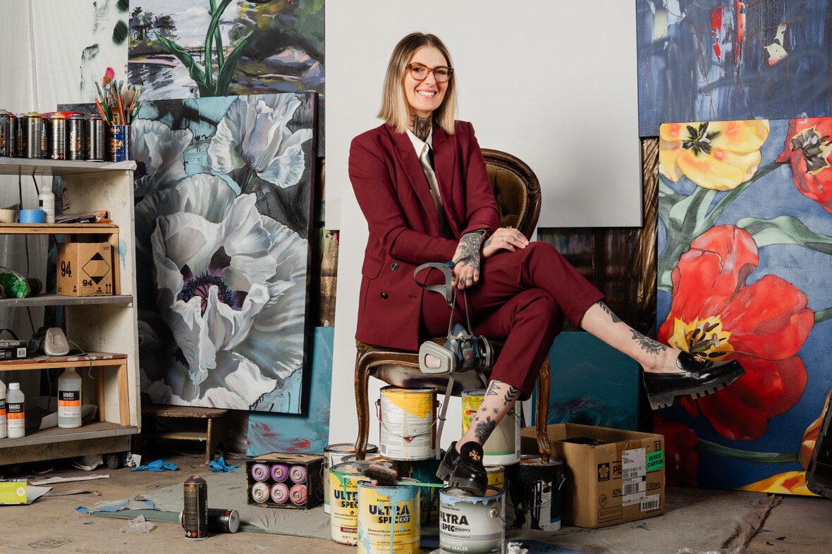 woman with blonde hair in maroon suit sitting in front of a few floral murals, with her foot on a paint can