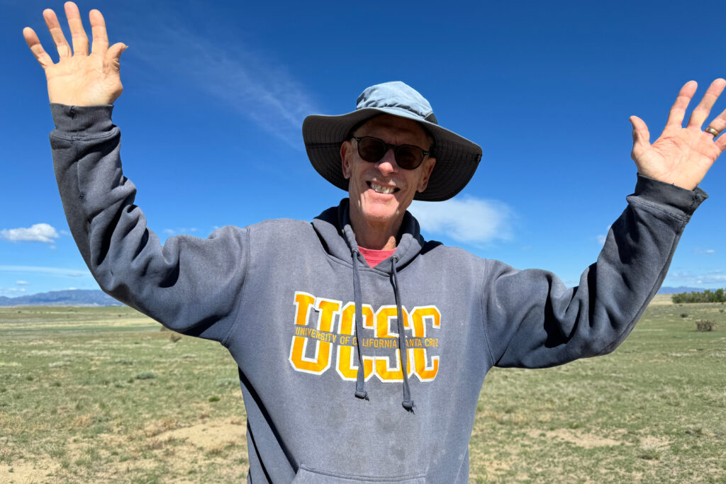 A man wearing a hat and a hoodie stands in a field with his hands raised.