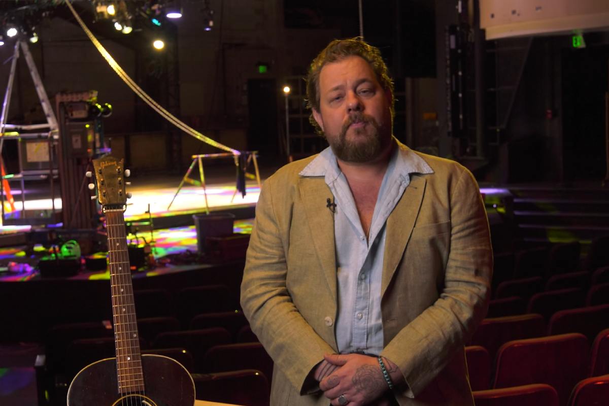 Man with beard standing next to a guitar in an auditorium, with a stage behind him.