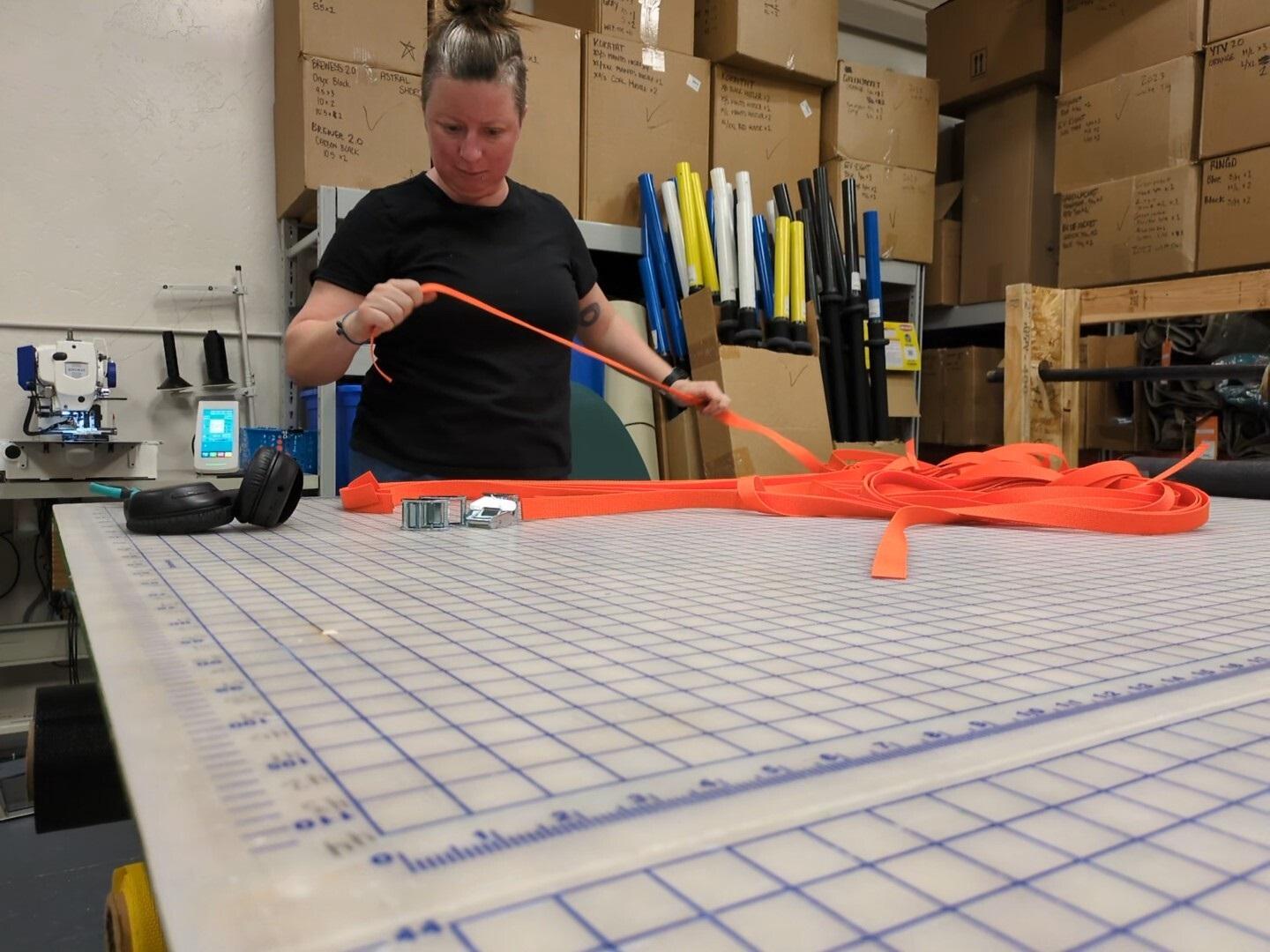 A woman is seen working on strips of orange straps. She is standing behind a table that has square measurements on it.