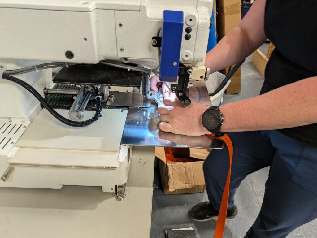 A woman's hands are seen setting an orange strap under a sewing machine to attach cam buckles for river rafting products.