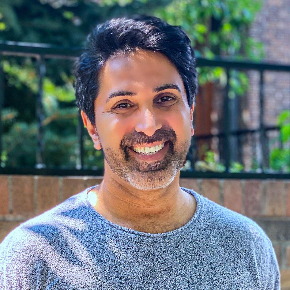 Man with black hair smiles at camera in the sun.