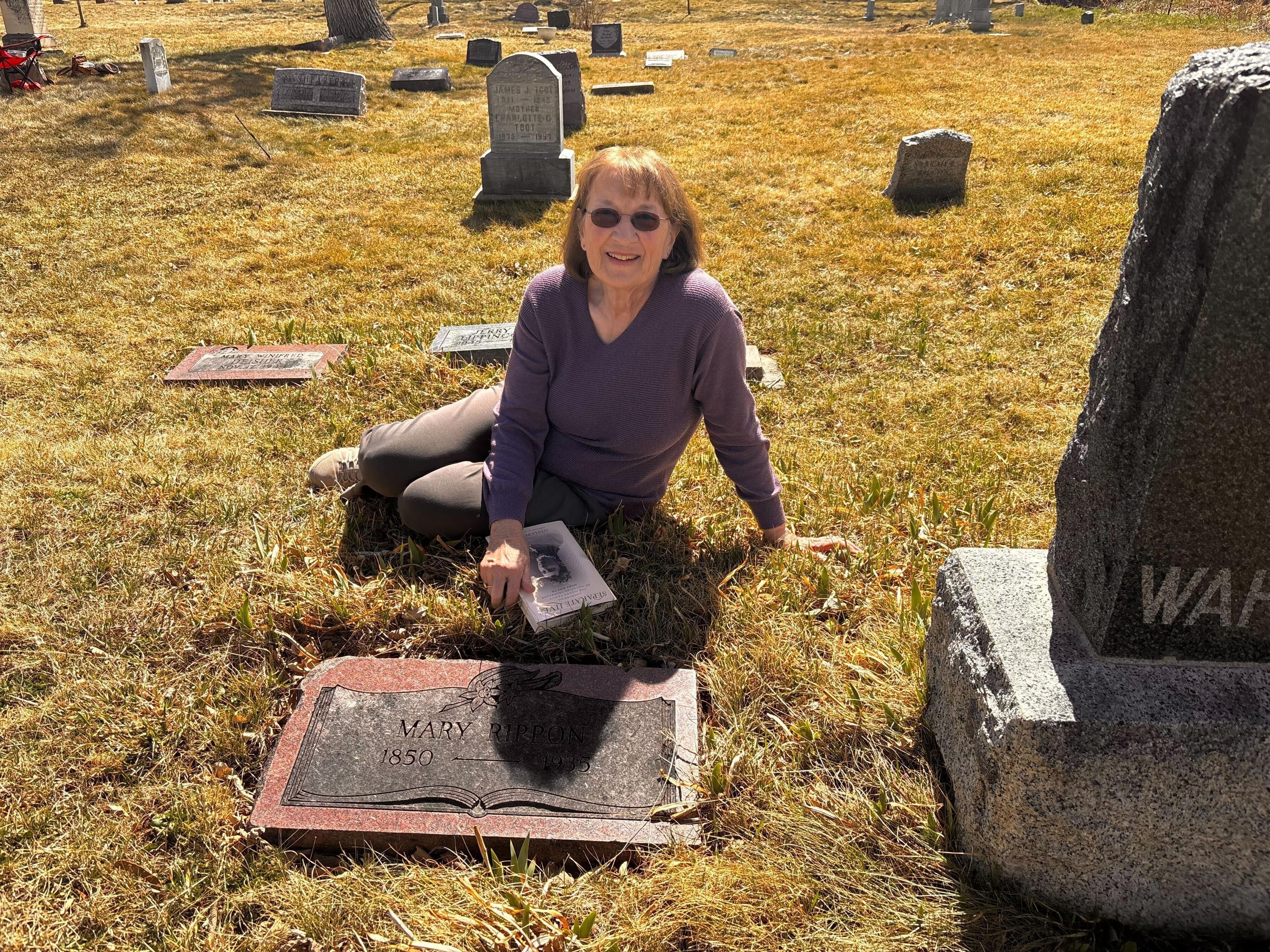 A woman is sitting on the ground next to a grave marker in a cemetery in Boulder.