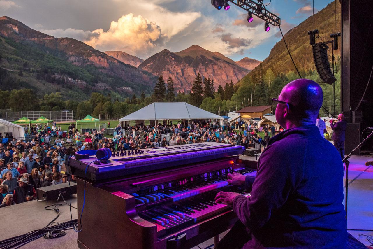 A man plays piano on stage, looking out over a lawn full of people in the beautiful box canyon of Telluride.