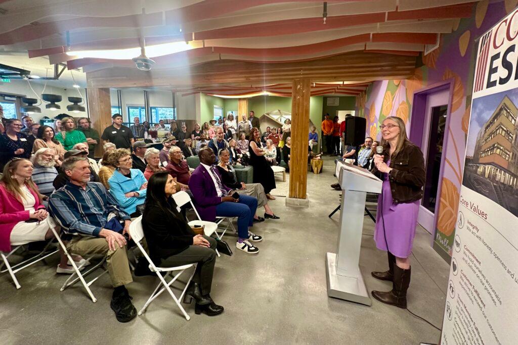 A woman in a purple dress speaks at a lectern to a room full of people sitting in folding chairs.