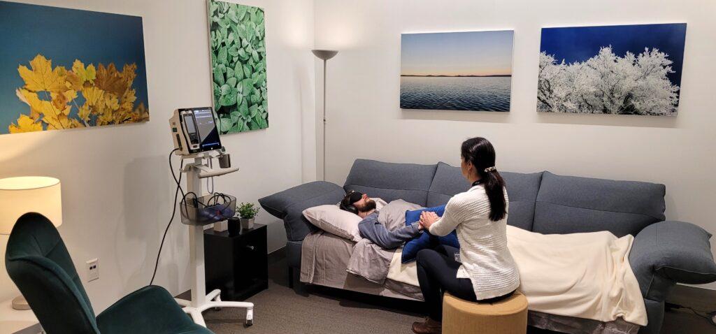 A facilitator holds the hand of a patient lying on a coach under blanket and an eye mask who is undergoing therapy.