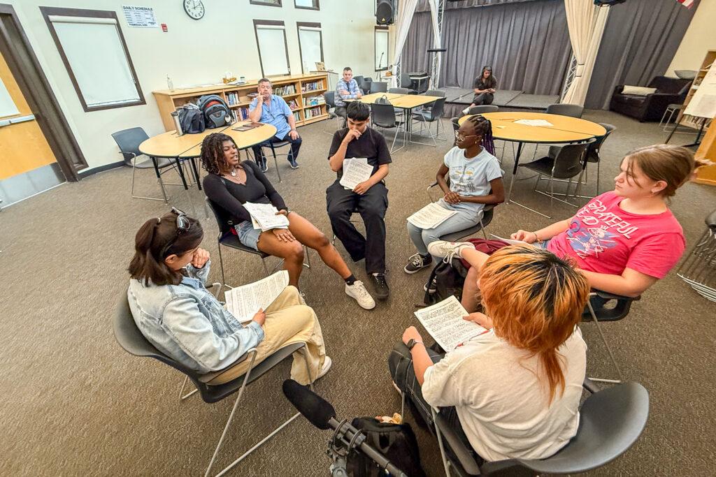 A group of six teenagers sit in a circle facing each other in a classroom. In the background, teachers listen to the discusiion.