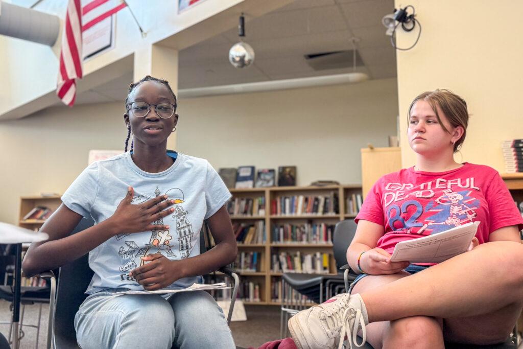 A Black teenage girl holds her right hand to her chest as she speaks in a classroom. A white teenage girl to her left looks and listens.