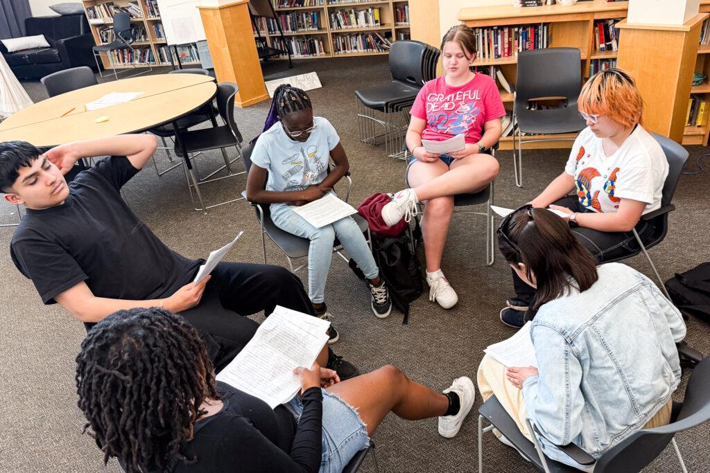 A group of six teenagers sit in chairs in a circle in a classroom. They each hold notes.
