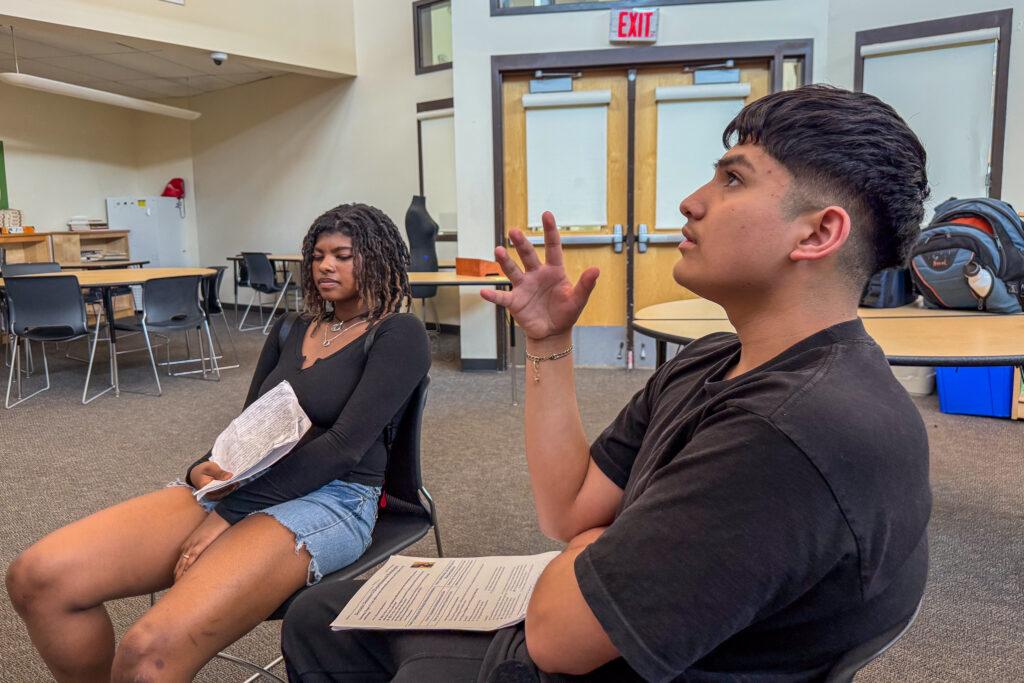 Side profile of a Latino teenage boy, who is holding his right hand up, palm facing the side, in a hand gesture as he speaks in a classroom. A Black teenage girl sits to his right listening. The are both holding notes.