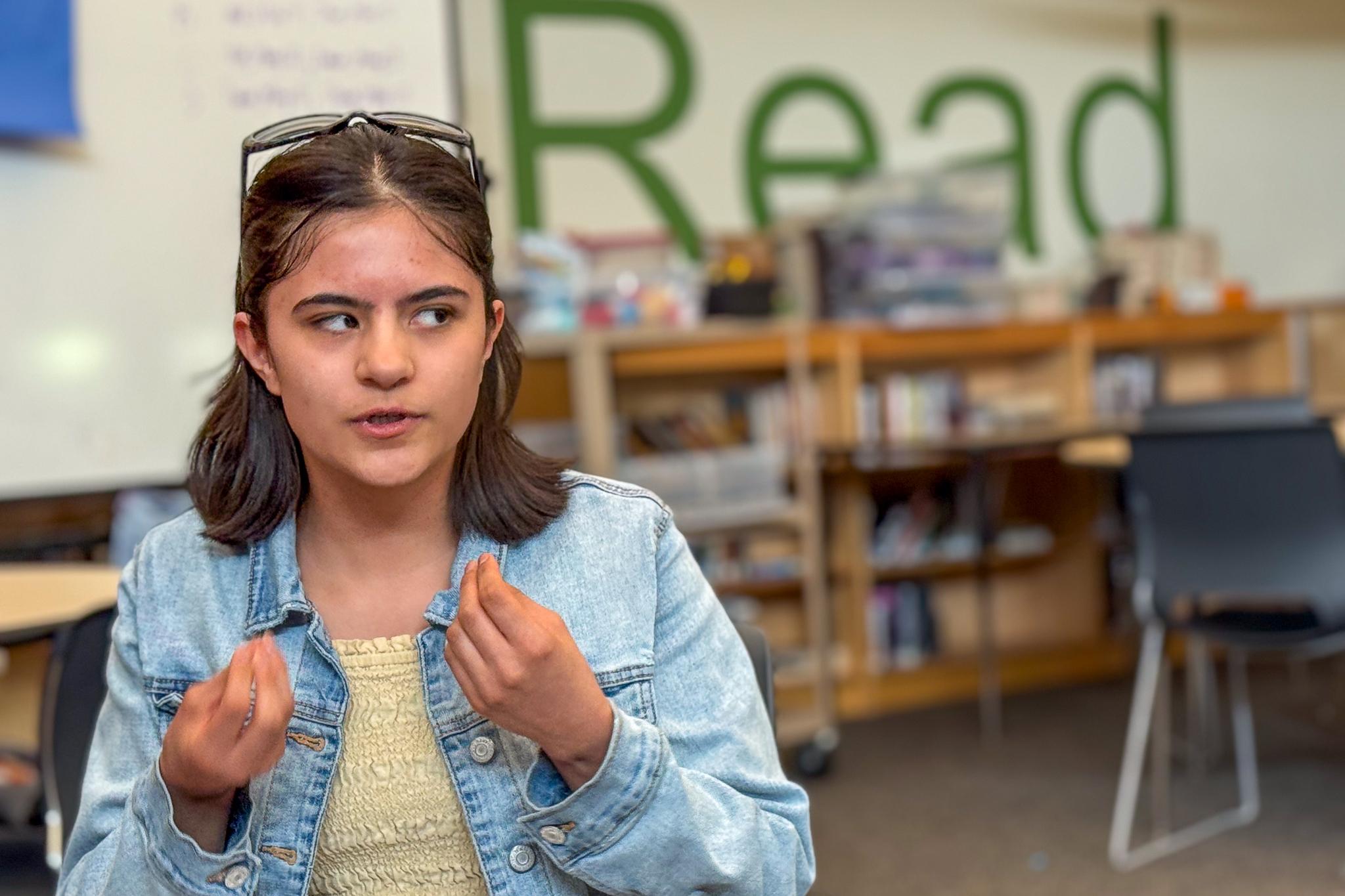 A teenage girl holds out her hands with her fingers pinches together, making a hand gesture as she speaks in a classroom. The wall behind her says "Read" in giant letters.