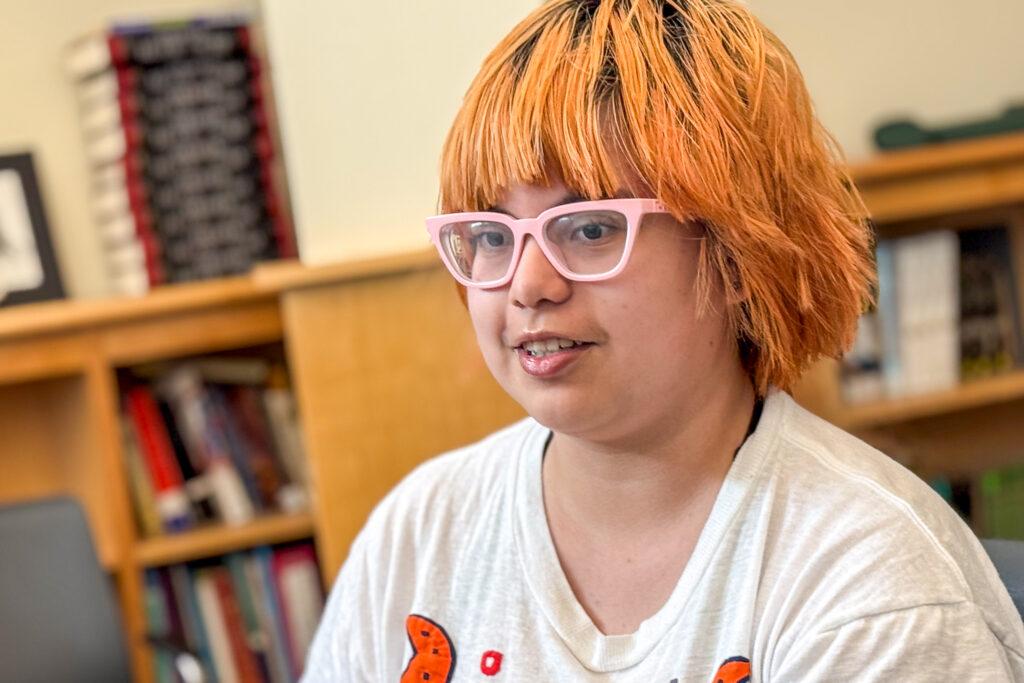 A portrait of a teenage student with dyed orange hair and pastel-pink-framed glasses sitting in a classroom.