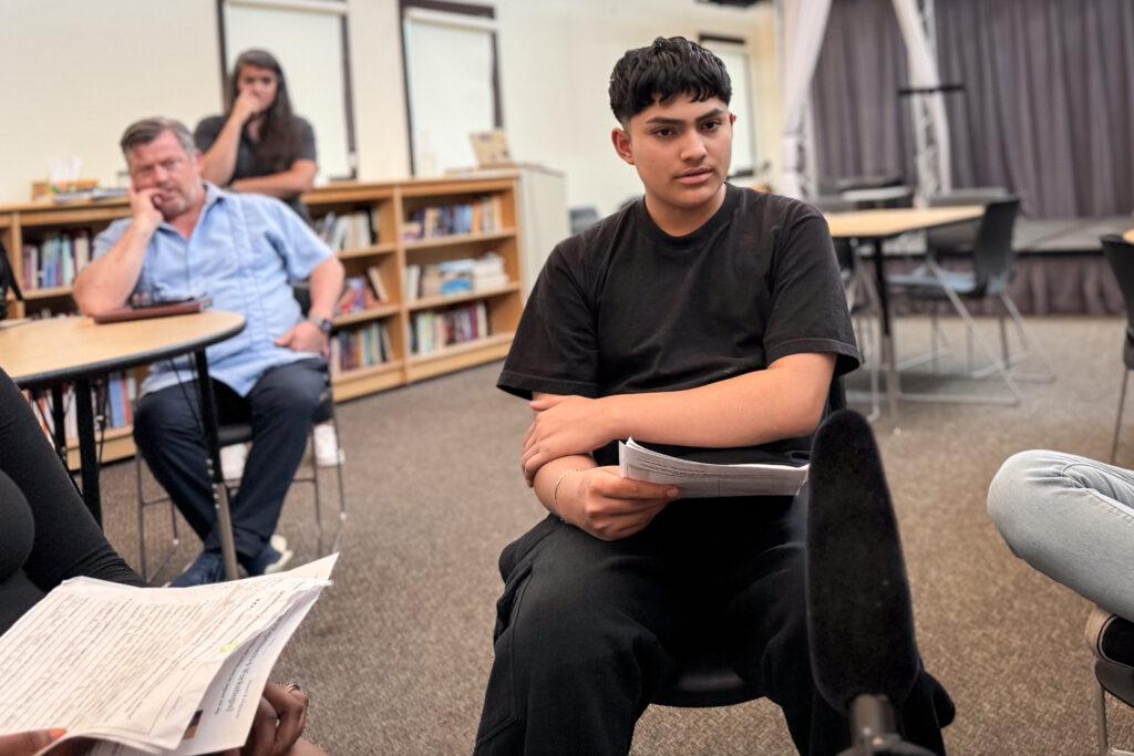 A teenager sits on a chair in a classroom holding papers. He is wearing all black and has one arm across his body.