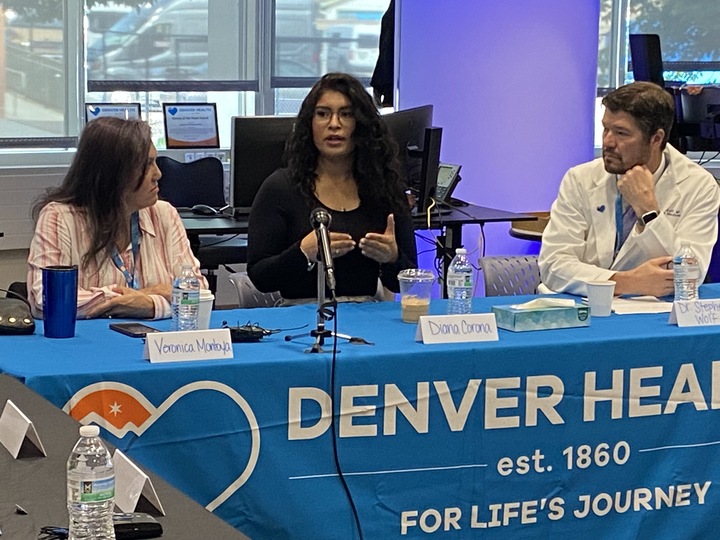 A Latino woman makesa gesture with her hand. pointing her fingers together with her palms facing her. She sits at a table with a blue banner that reads Denver Health. A doctor in a white coat listens on her left as a woman on her right also listens.