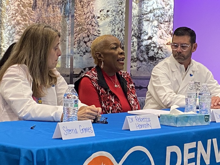 An okder Black woman sits at a conference table with a blue banner that reads Denver Health and speaks while a woman doctor on her left and a male doctor wearing glasses on her right, both wearing white coats, look on