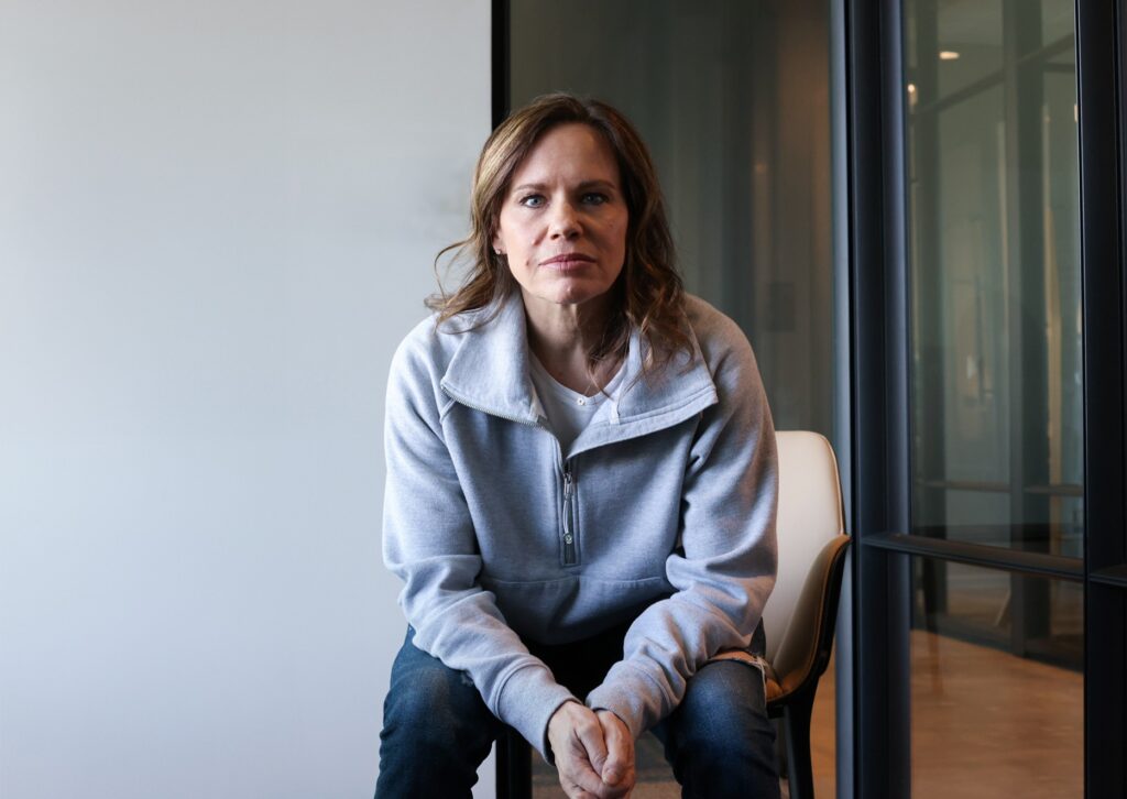 A woman with light brown hair, wearing a gray zip-up jacket and jeans, sits on a chair in a modern office space, looking directly at the camera with a serious expression.