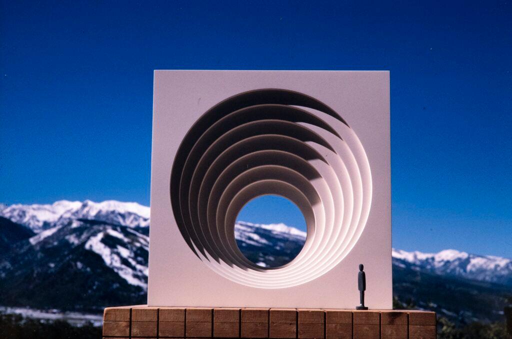 A model of a square, white gate with receeding circles inside. A small human figure stands next to the gate, showing how large it is.