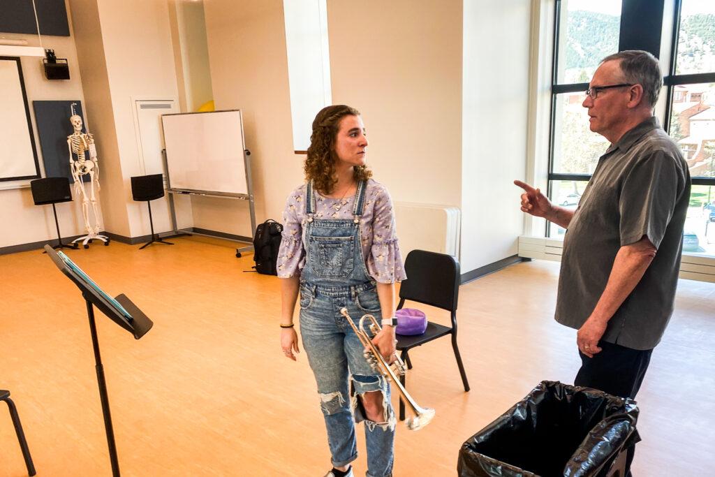 A woman in overall with rips in the knees stands looking at her teacher while she holds a trumpet in her resting hands. The teacher is an older man werea a gray short-sleeved button-up short and black framed eyeglasses. They are in a classroom.
