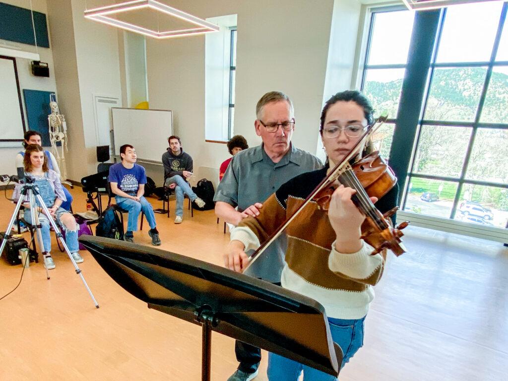 A older man teacher has his hand on the elbow of a student as she plays the viola in a classroom.