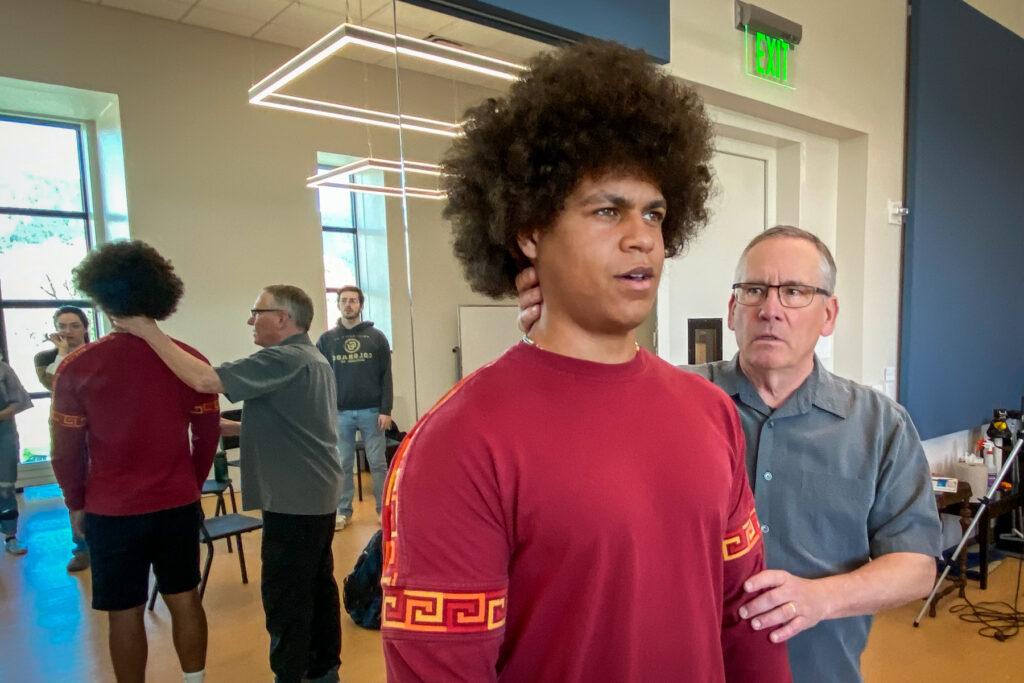 An older man in a gray button-up shirt holds the back of a Black student's neck. The student has an afro and is wearing a red shirt. In the background, other students are reflected in the mirror behind the two men.