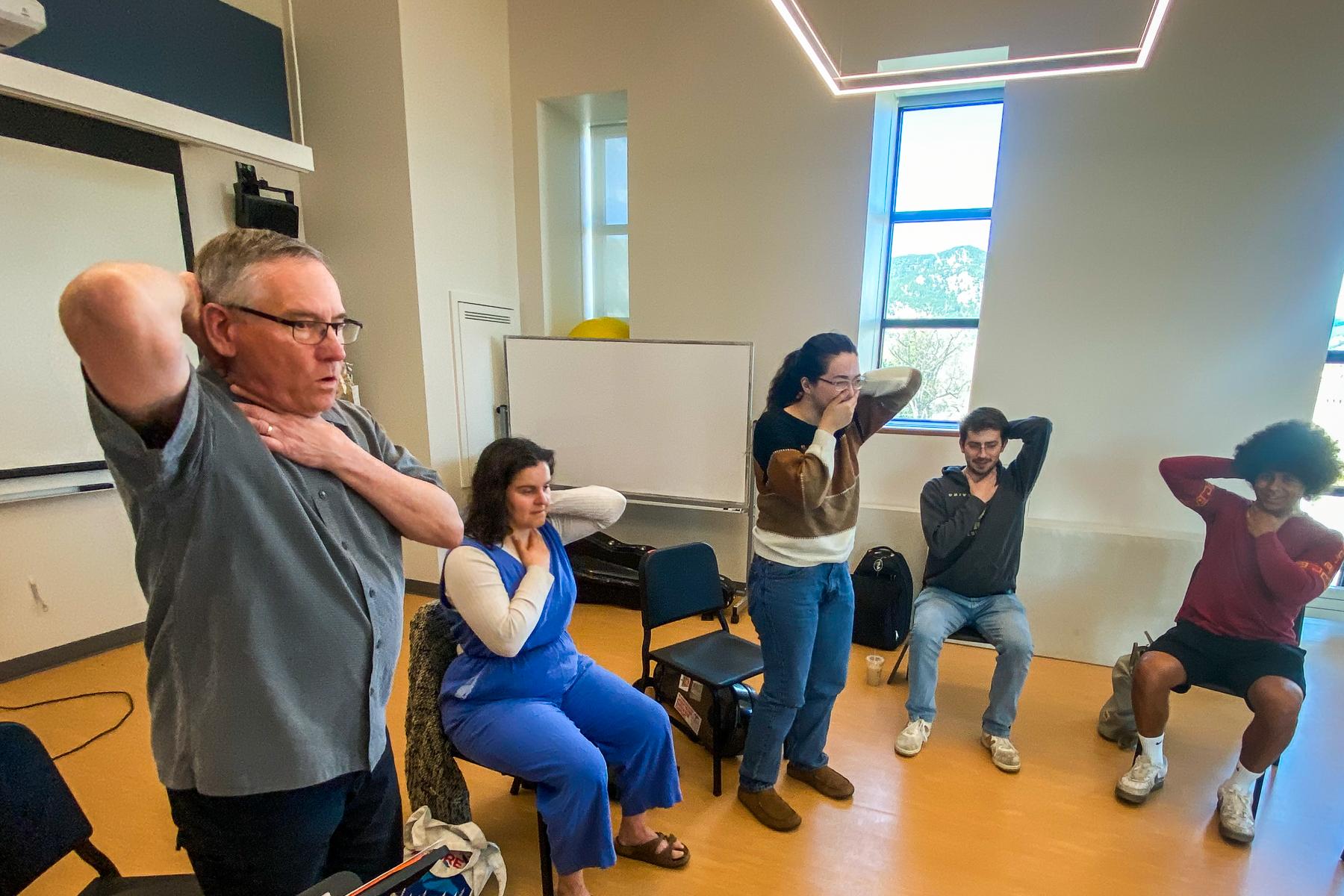 An older man in the forground has one hand on the back of his head on one at the base of his throat. Four younger people copy the position.