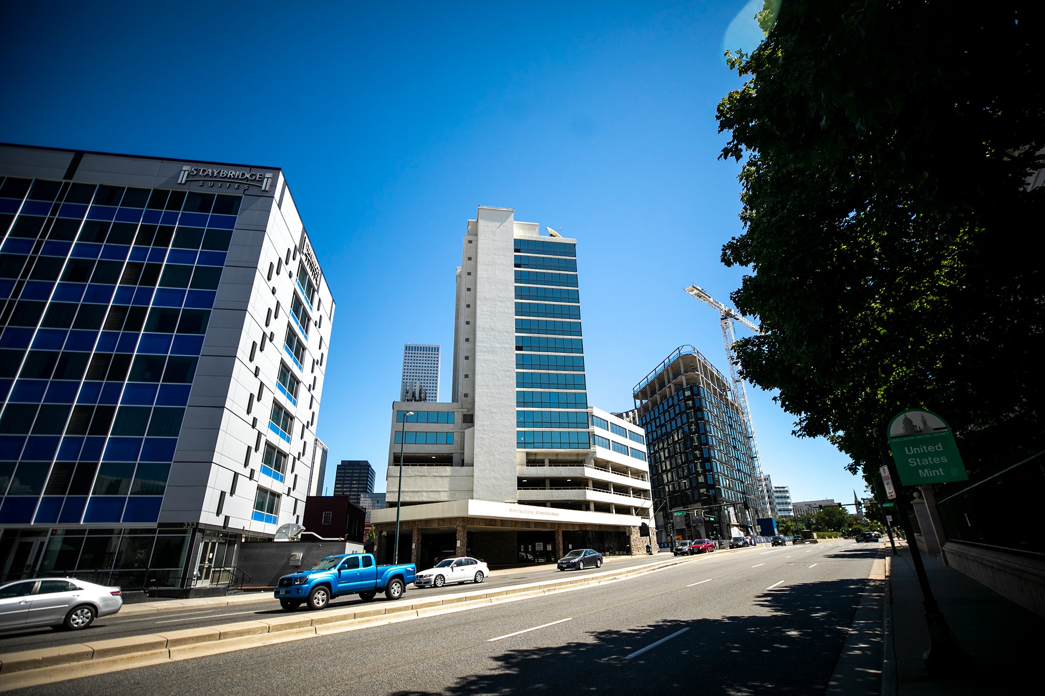 The image features a city street with tall buildings on both sides. There are several cars parked along the street, and a truck is visible on the left side. The scene is set on a sunny day, with the sunlight creating a bright atmosphere.