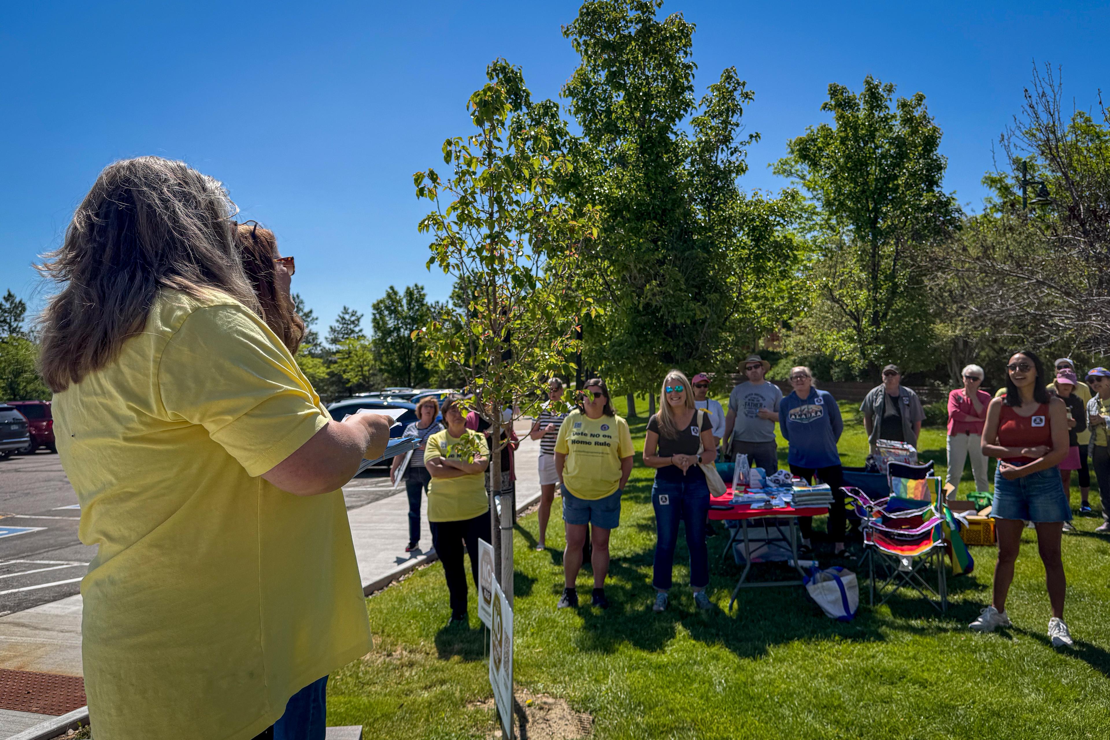 Two women in yellow shirts stand in the foreground addressing a small crowd of people standing on a lawn under sunny skies