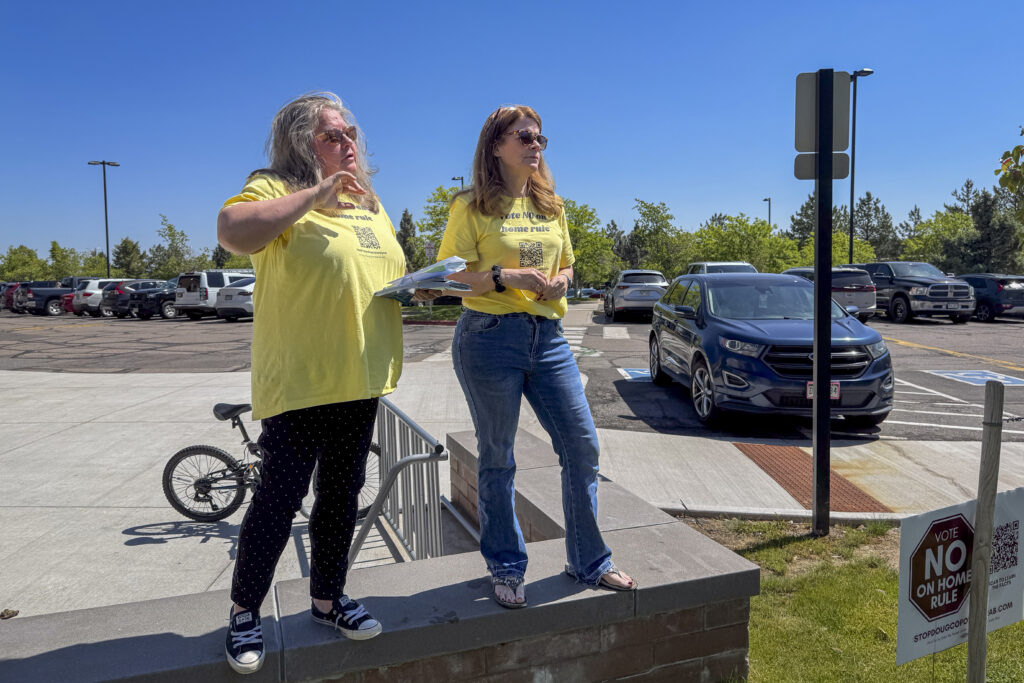 Two women in yellow shirts stand on a low wall with a parking lot behind them