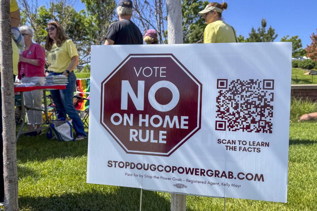 a large lawn sign in the foreground, in front of a table with people gathered around it, all outside on a sunny day