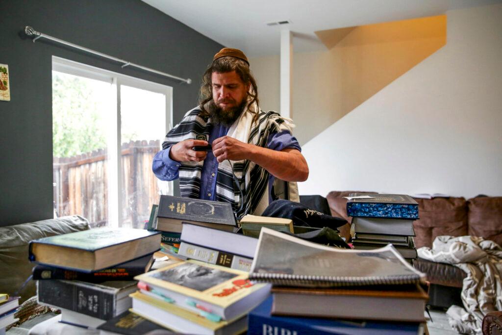 A man in traditional Jewish clothing is standing behind a collection of books written in Hebrew.